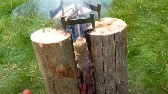 На лужайке установлена конструкция из трех бревен. Они горят внутри и запекают овощи на шампуре